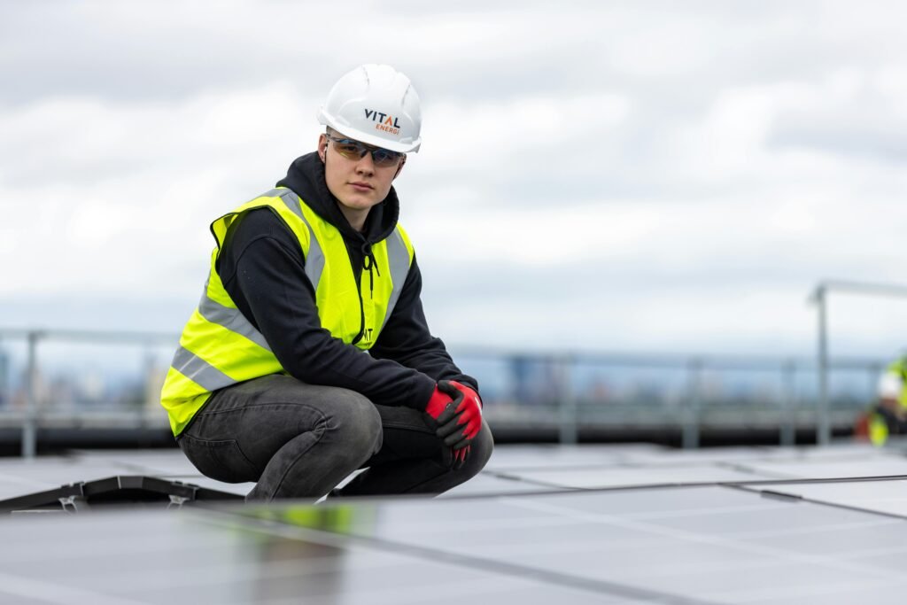 A construction worker inspects rooftop solar panels, wearing safety gear and high visibility vest.