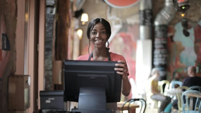 Positive young woman in uniform smiling while standing at counter desk in cafe